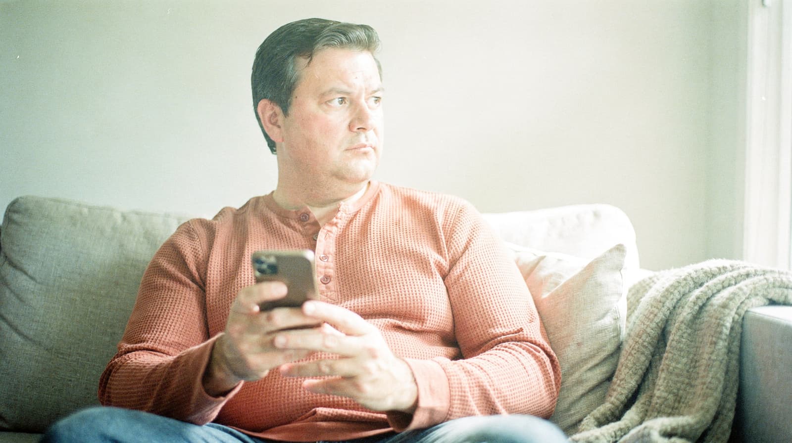A man in a red sweater sits on a couch holding a smartphone, looking out a window.