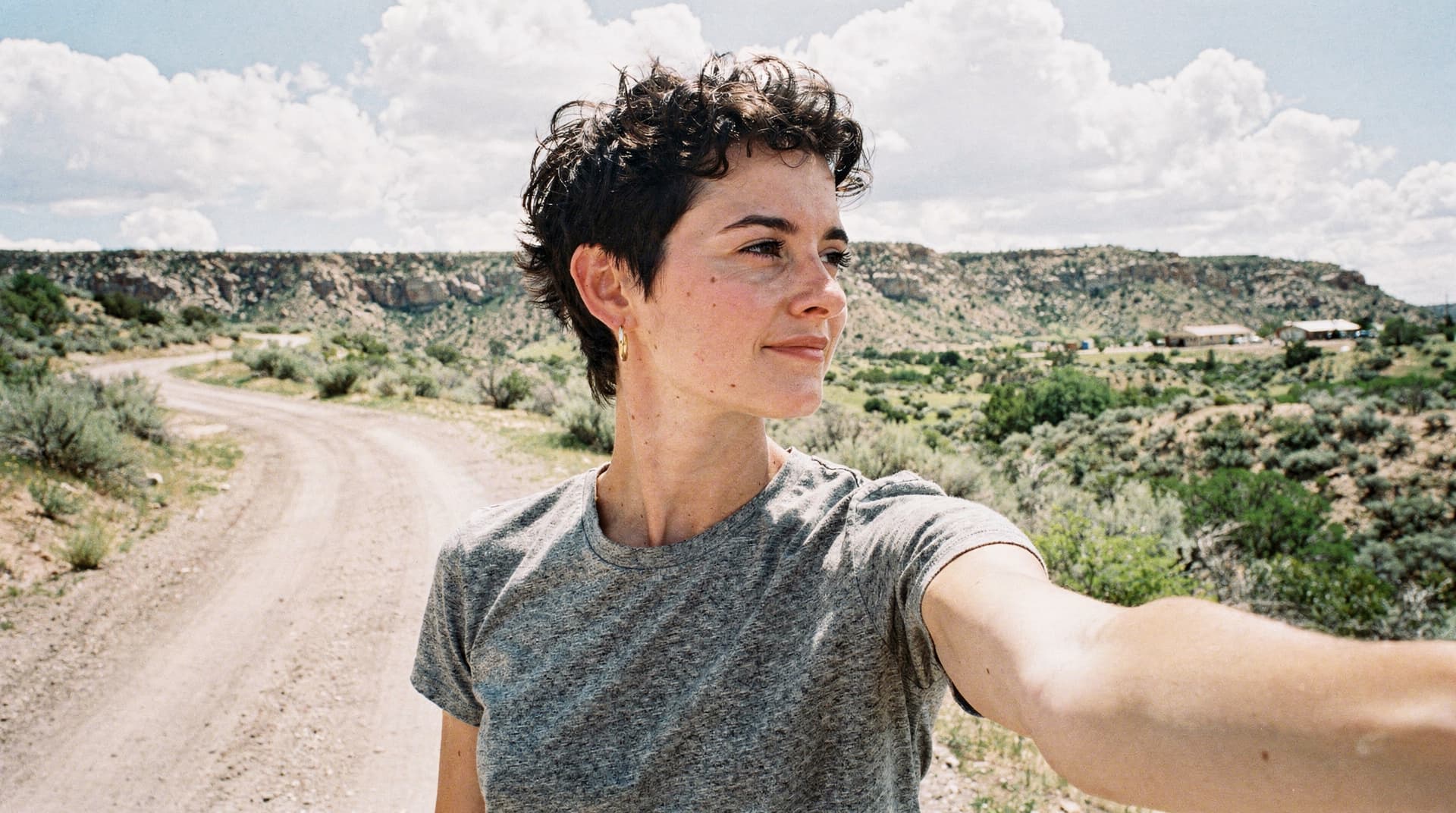 A person with short hair takes a selfie on a dirt road in a desert landscape with clouds in the sky.