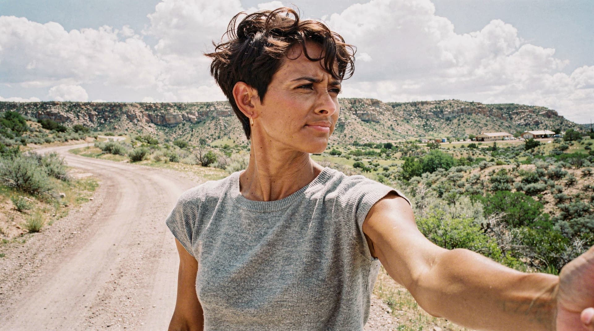 A person with short hair takes a selfie on a dirt road in a desert landscape with clouds in the sky.