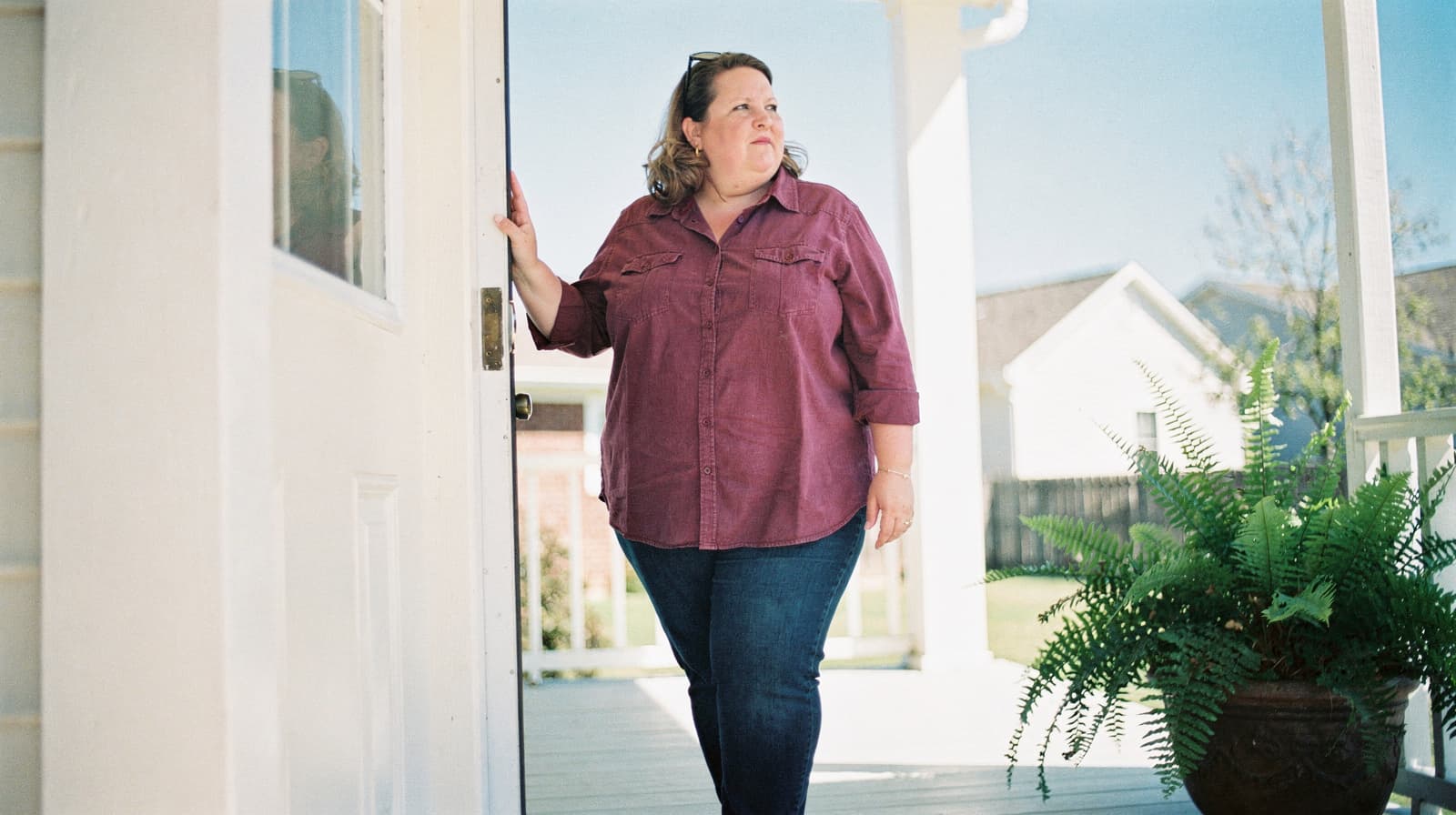 A woman in a purple shirt stands on a porch, holding a door open, with a potted fern nearby.