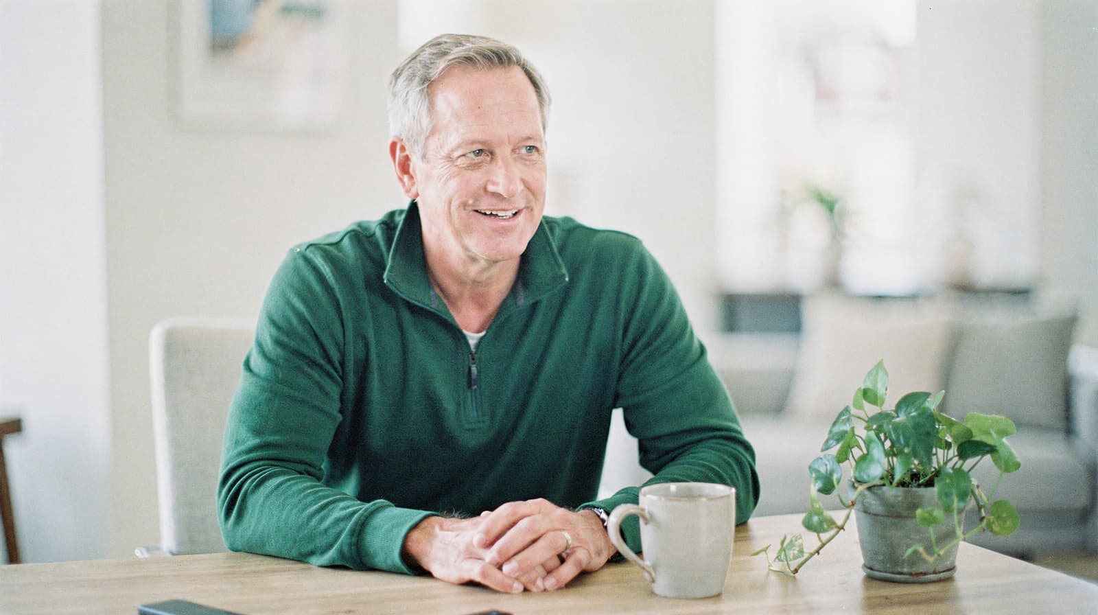 A man in a green sweater sits at a table with a mug and a potted plant indoors.