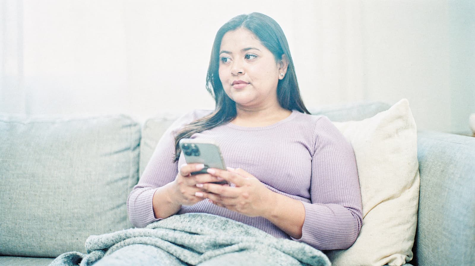 A woman sits on a couch holding a smartphone, wrapped in a blanket.