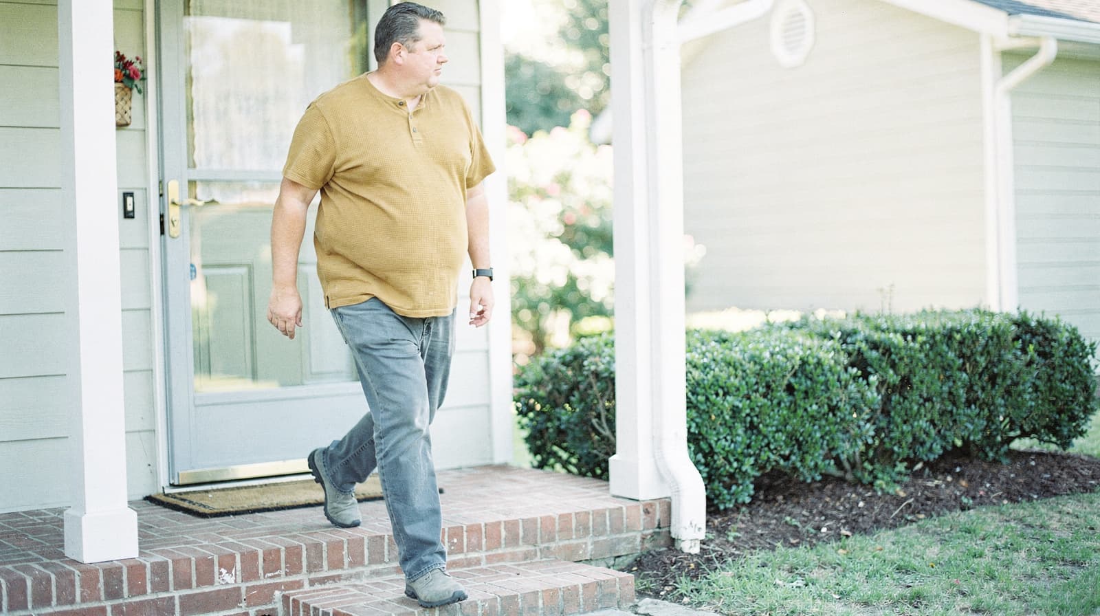 A man in a yellow shirt steps off a brick porch in front of a house with white columns and green shrubs.