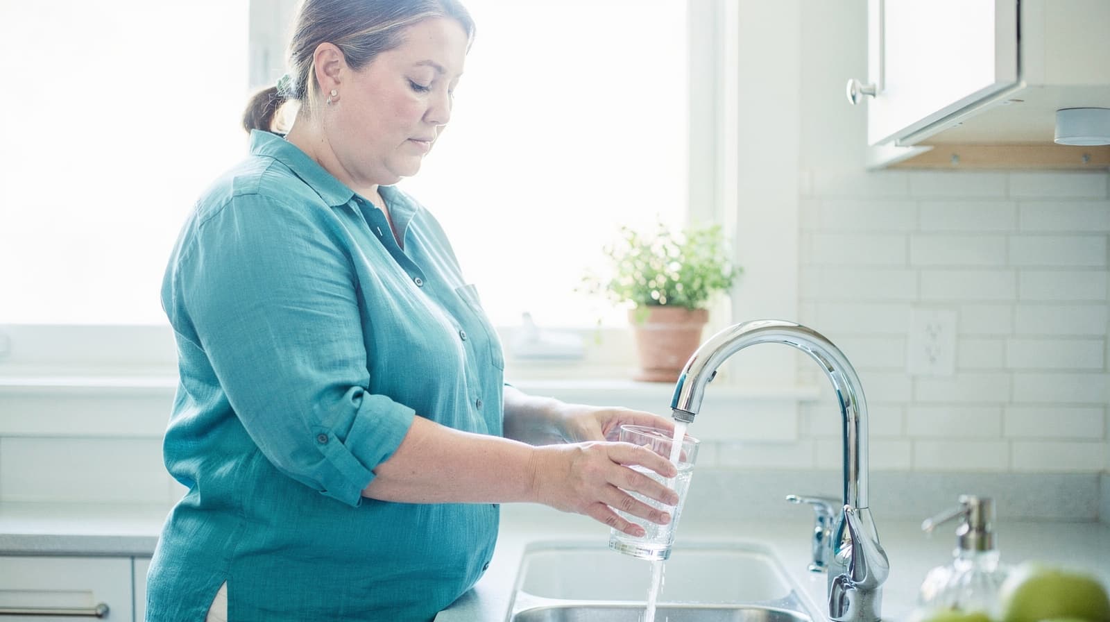 A woman in a teal shirt fills a glass with water at a kitchen sink.