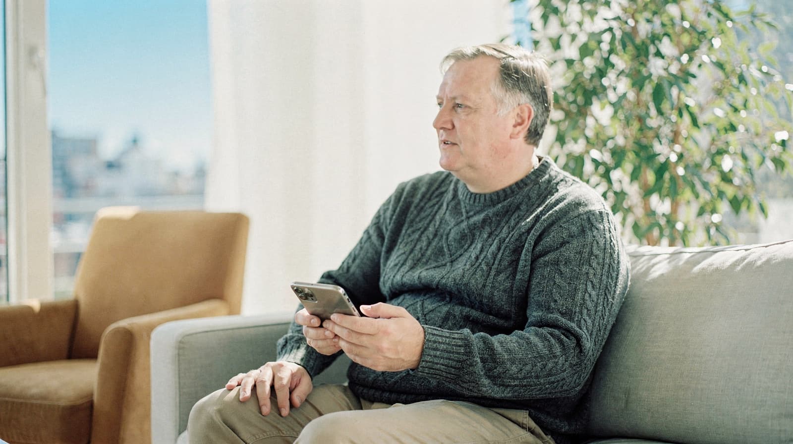 A man sits on a couch holding a smartphone, with a large plant and an armchair in the background.