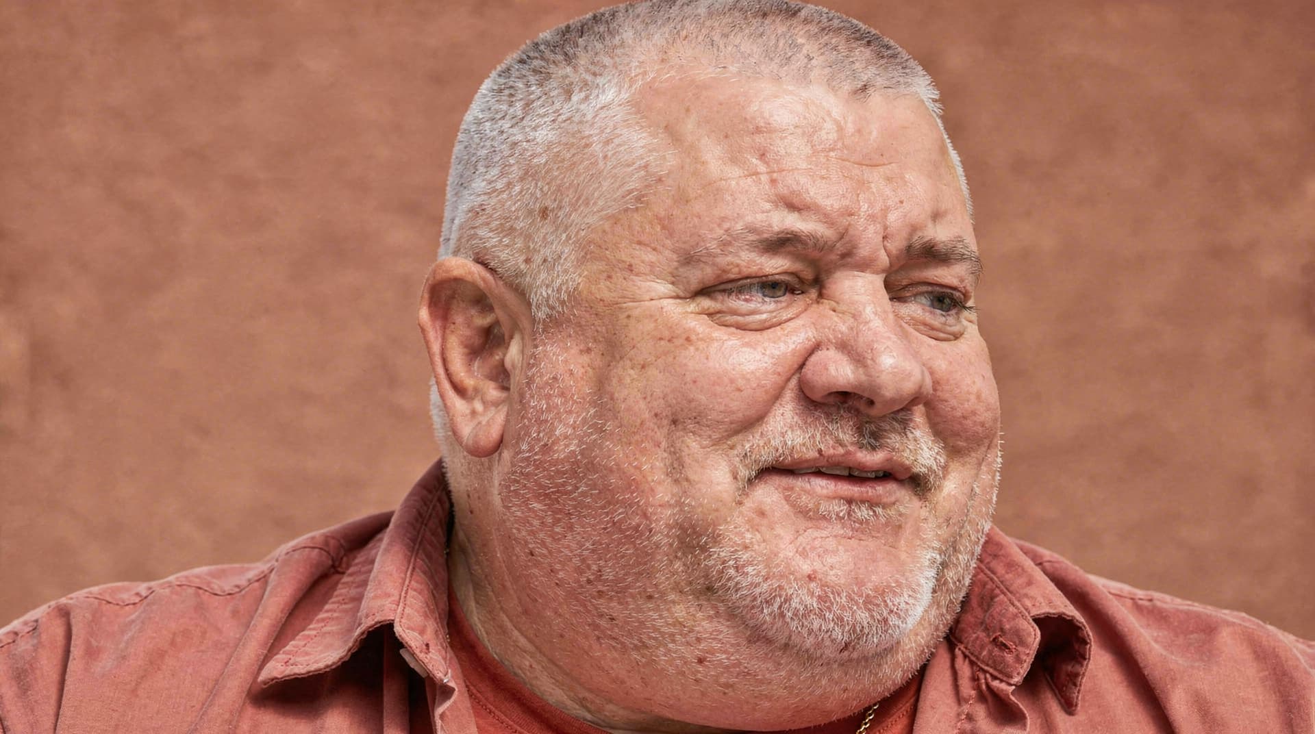 A man with short hair and a beard smiles while looking to the side against a plain background.