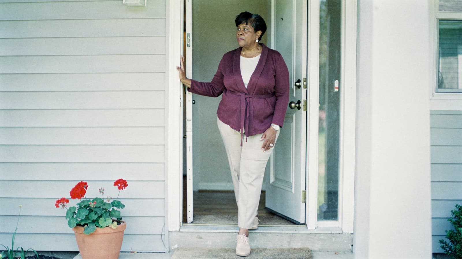 A woman stands in a doorway of a house, looking to the side, with a potted plant nearby.