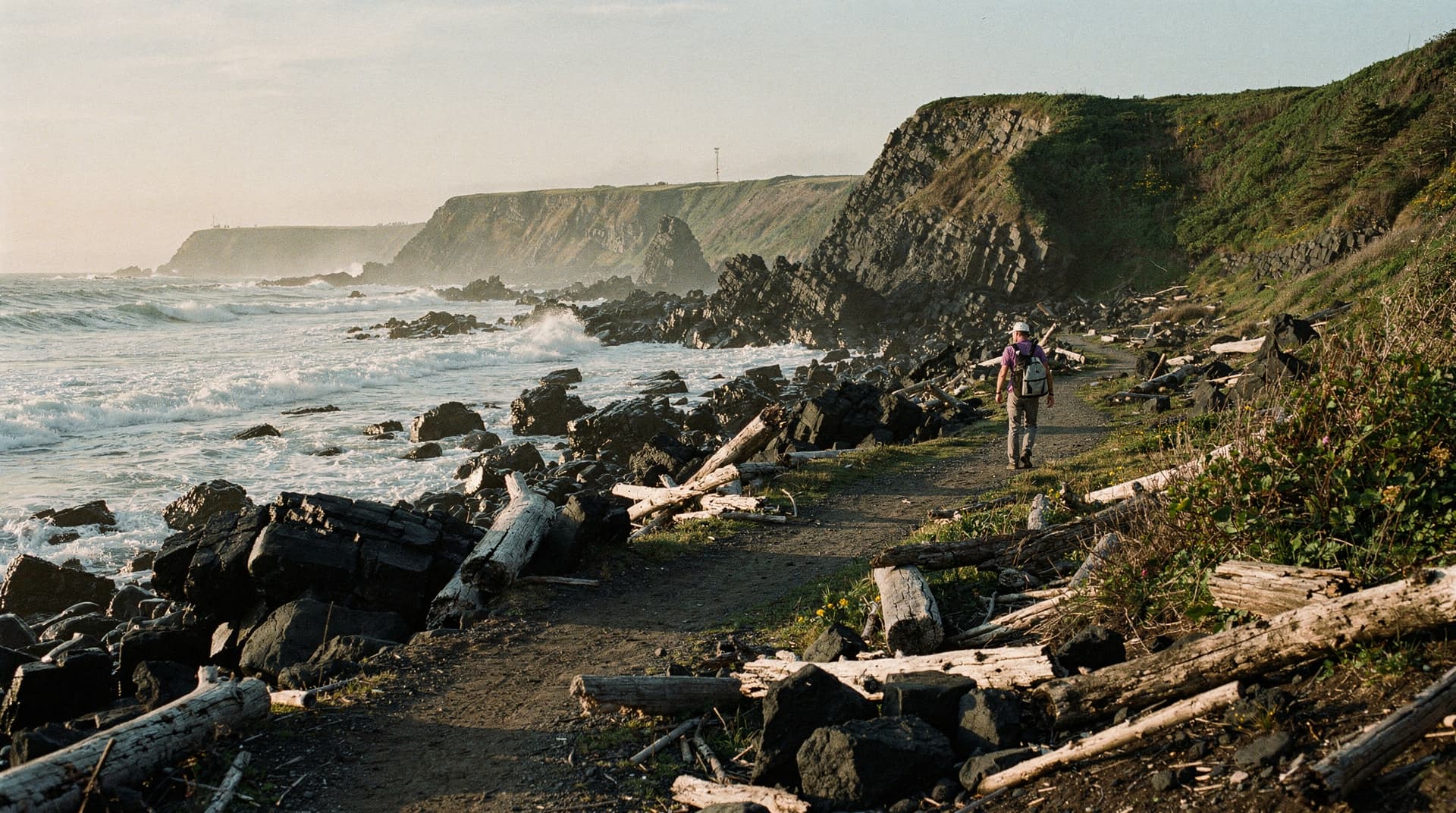 A person walks along a rocky coastal path with waves crashing against the shore and cliffs in the background.