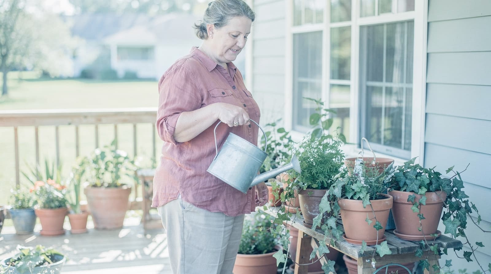 A woman waters potted plants on a wooden deck outside a house.