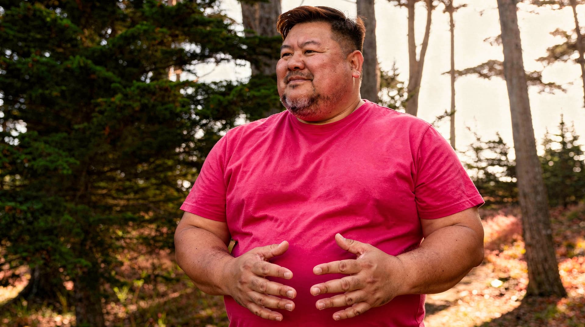 A man in a red shirt stands in a forested area with sunlight filtering through the trees.