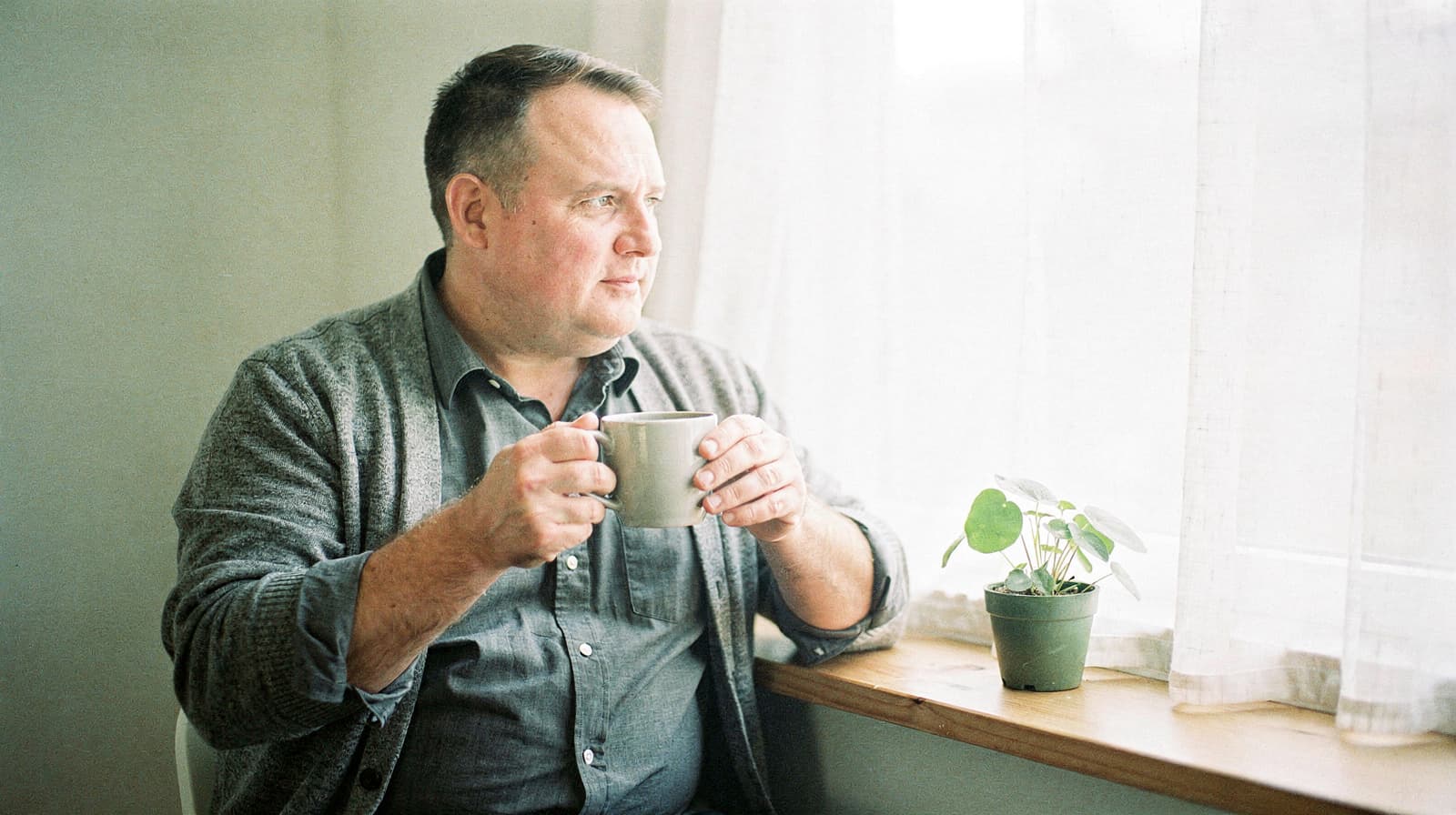 A man holding a mug looks out a window while sitting next to a small potted plant.