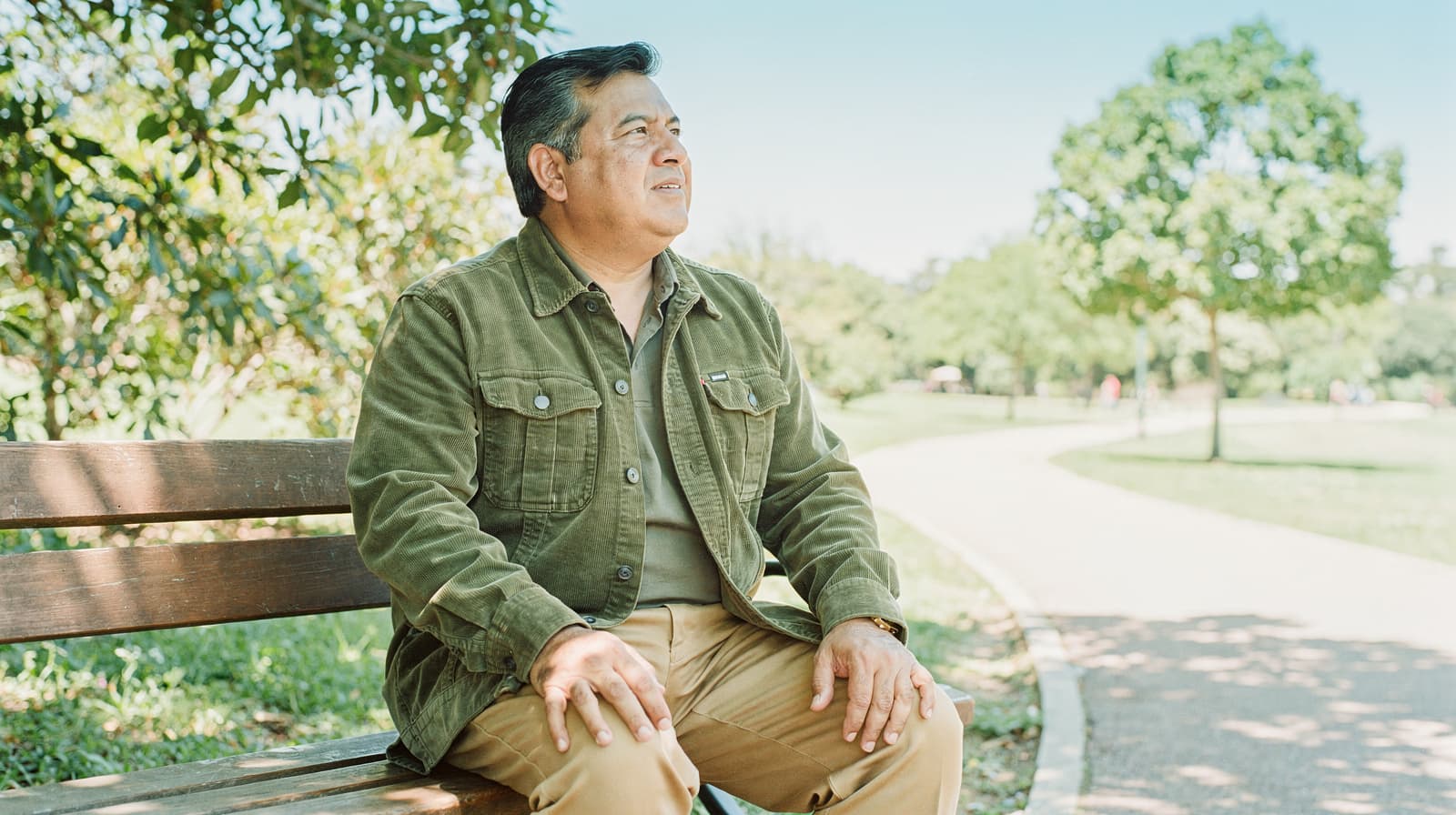 A man sits on a wooden bench in a park, looking into the distance on a sunny day.