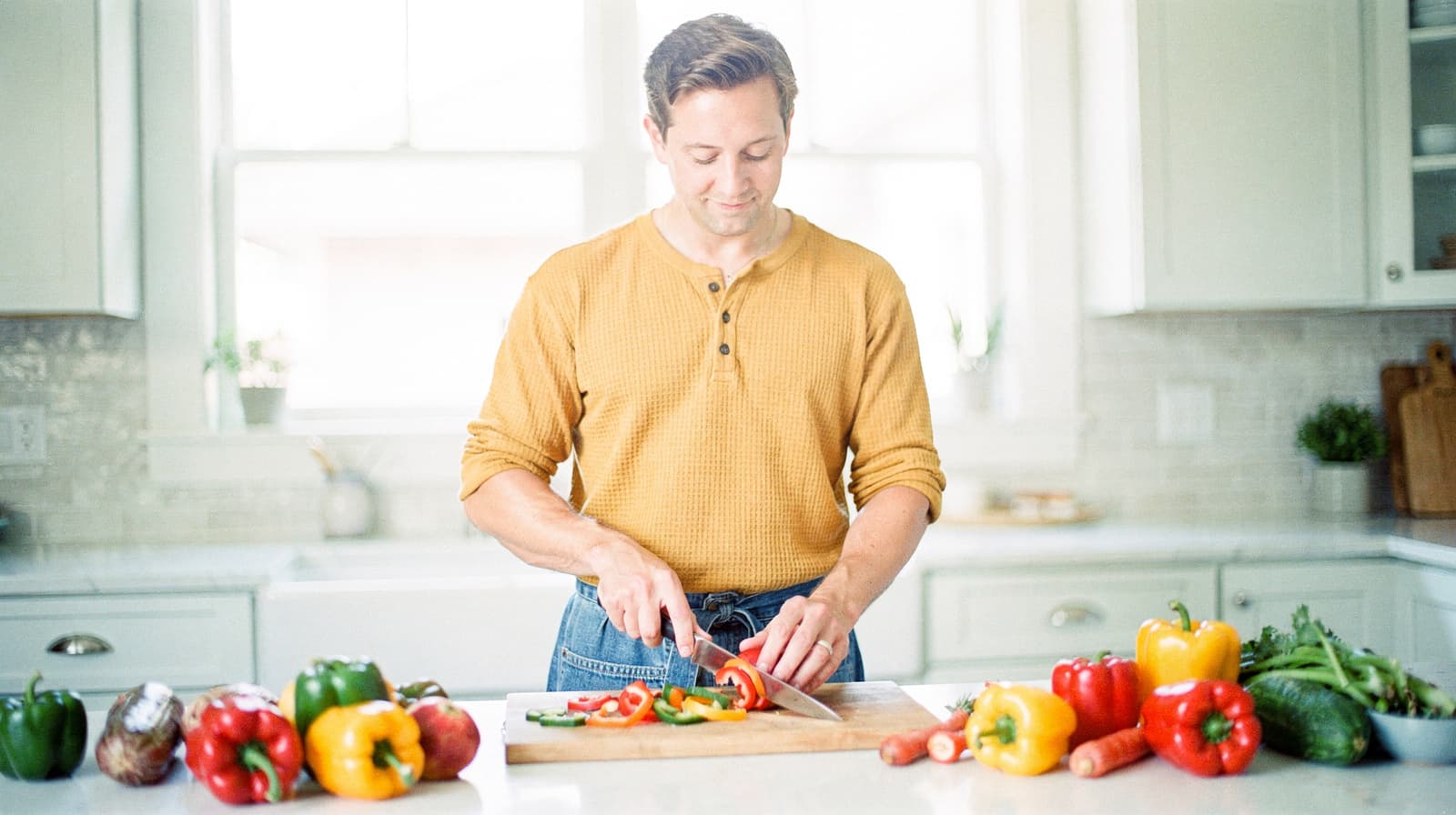 A man in a yellow shirt slices vegetables on a wooden cutting board in a bright kitchen.