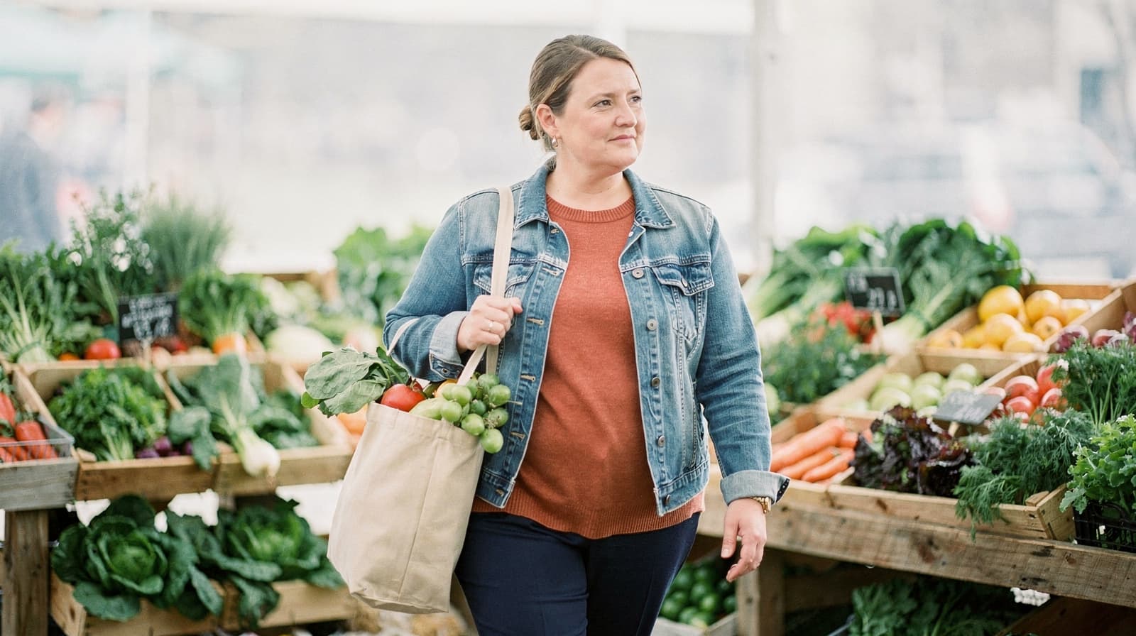 A woman carries a tote bag filled with vegetables while walking through a farmers market.