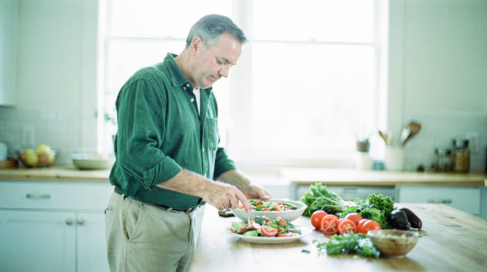 A man in a green shirt prepares a salad in a bright kitchen with fresh vegetables on the counter.