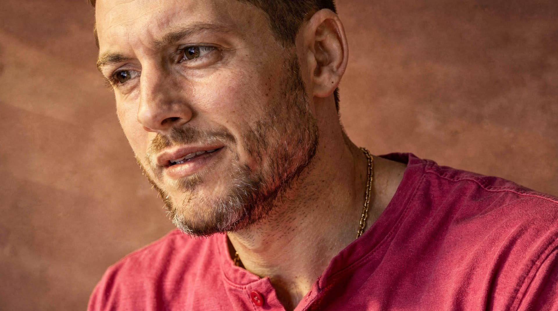 A man with a beard and short hair wears a red shirt and a gold chain, looking to the side against a brown background.