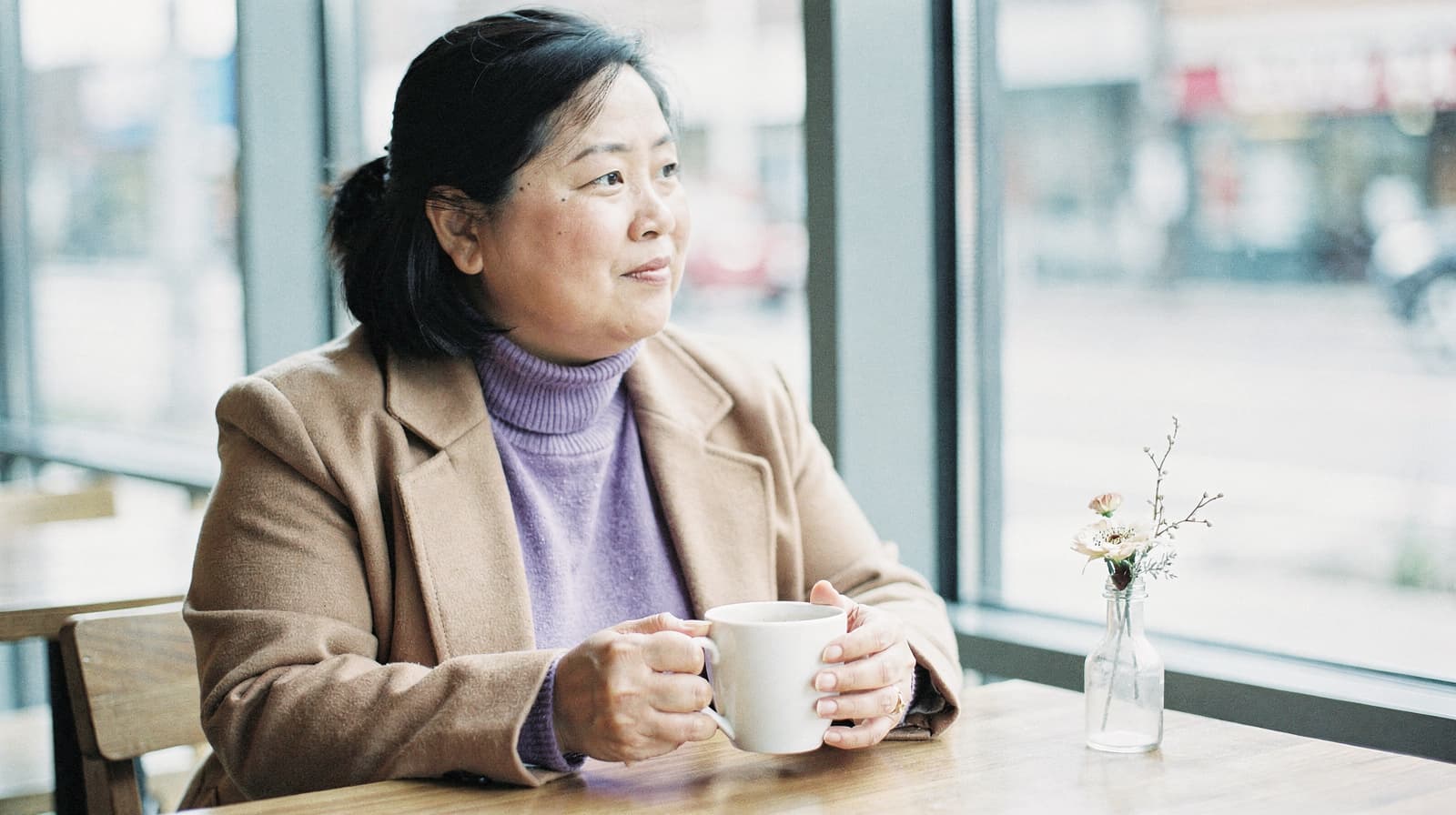 A woman sits at a café table holding a mug, looking out the window with a small vase of flowers beside her.