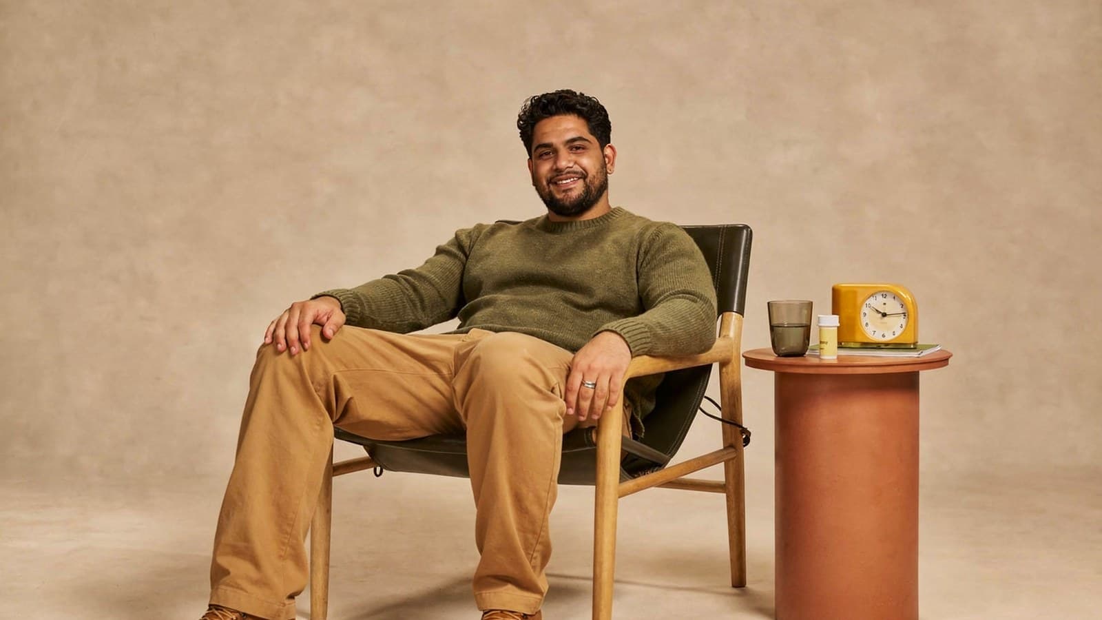 A man in a green sweater sits on a chair next to a small table with a clock and a cup.