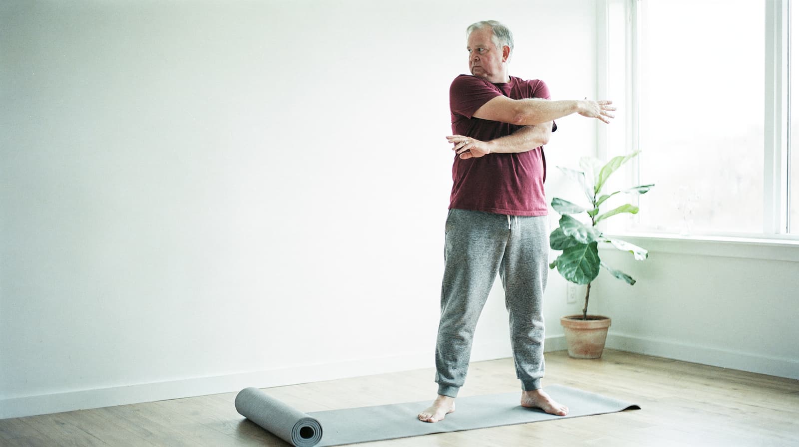 A man stretches on a yoga mat in a bright room with a potted plant by the window.