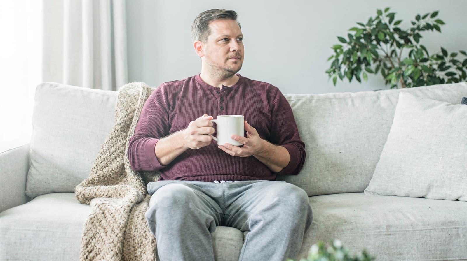 A man sits on a couch holding a mug, with a blanket draped beside him and a plant in the background.