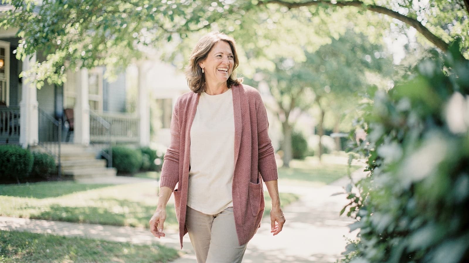 A woman walks along a sunny residential street lined with trees and houses.