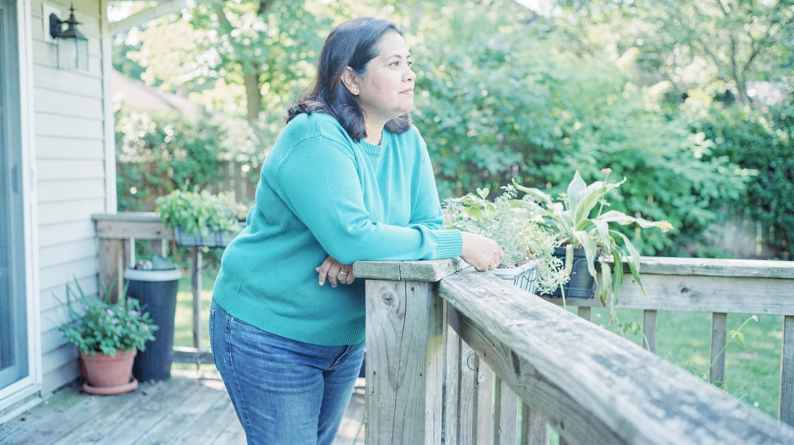 A woman in a blue sweater leans on a wooden deck railing, looking out at a garden.