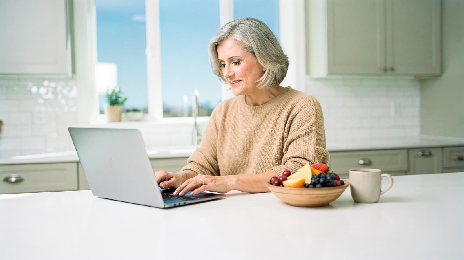 A woman with gray hair types on a laptop at a kitchen counter with a bowl of fruit and a mug nearby.