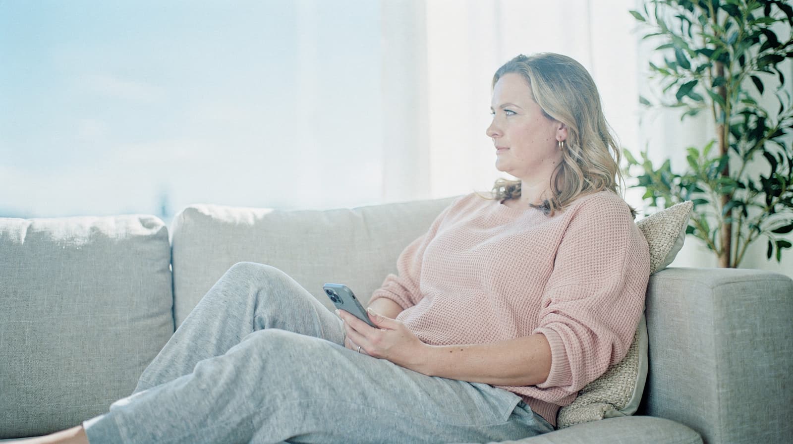 A woman in a pink sweater sits on a couch holding a smartphone, looking out a window with a plant nearby.