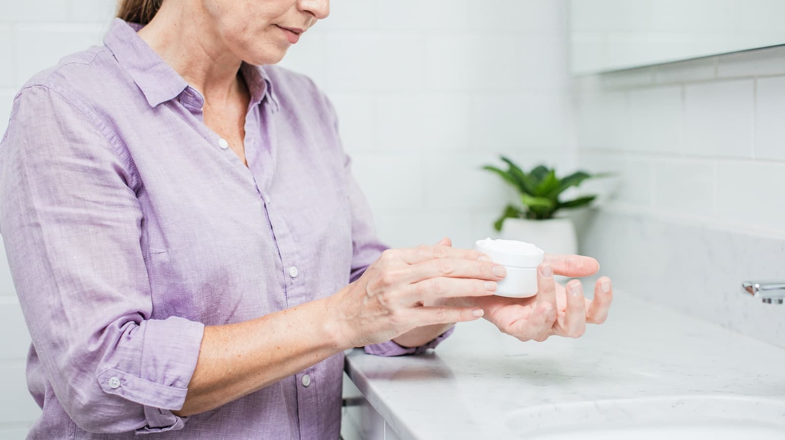 A woman in a lavender shirt applies cream from a small container in a white-tiled bathroom.
