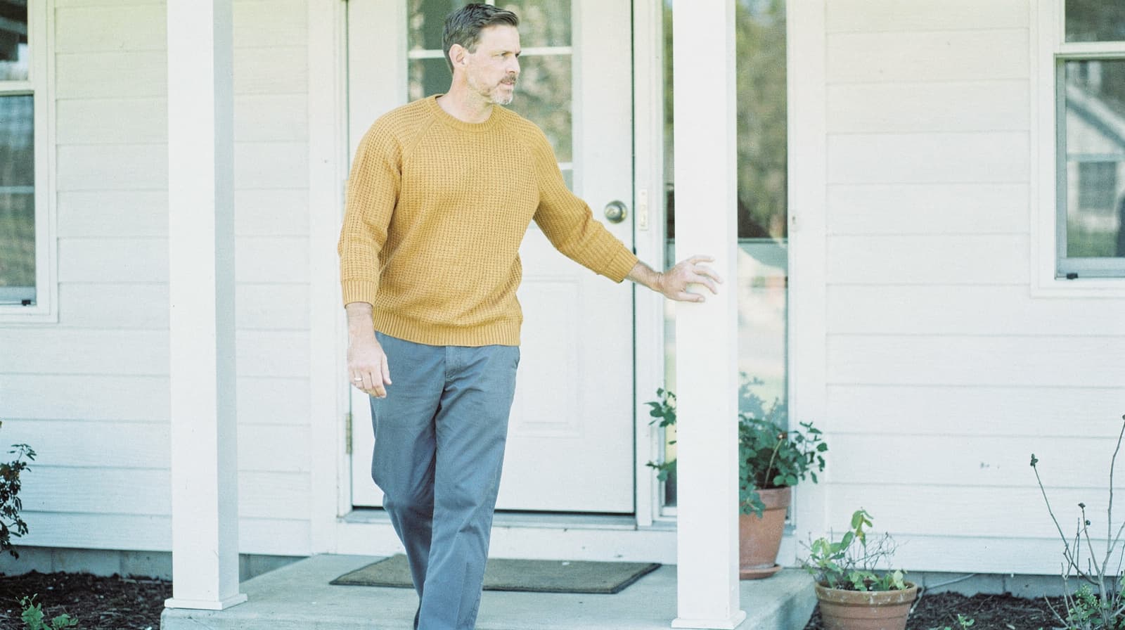 A man in a sweater stands on a porch, holding a column, with potted plants nearby.
