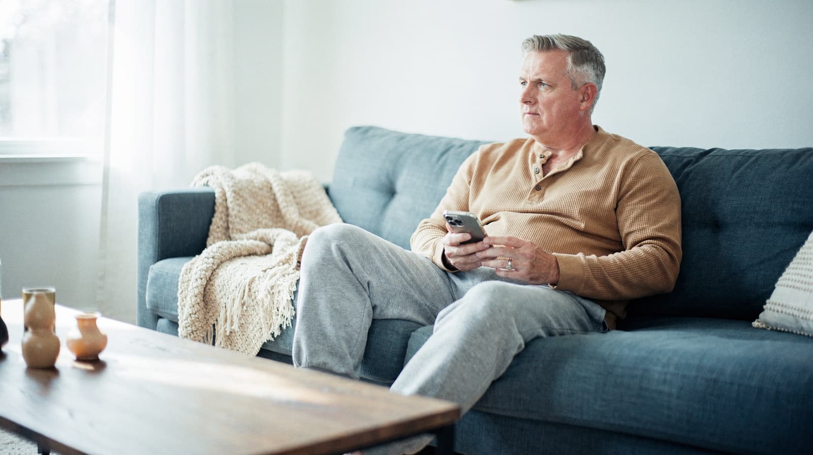 A man sits on a blue sofa holding a smartphone in a bright living room.