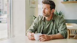 A man sits at a table in a cafe holding a mug and looking out the window.