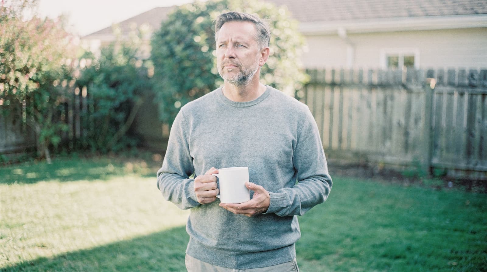 A man holding a mug stands in a backyard with grass and a wooden fence.