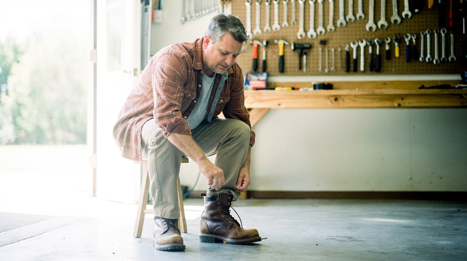 A man sits on a stool in a garage tying his boot laces, with tools hanging on a pegboard in the background.