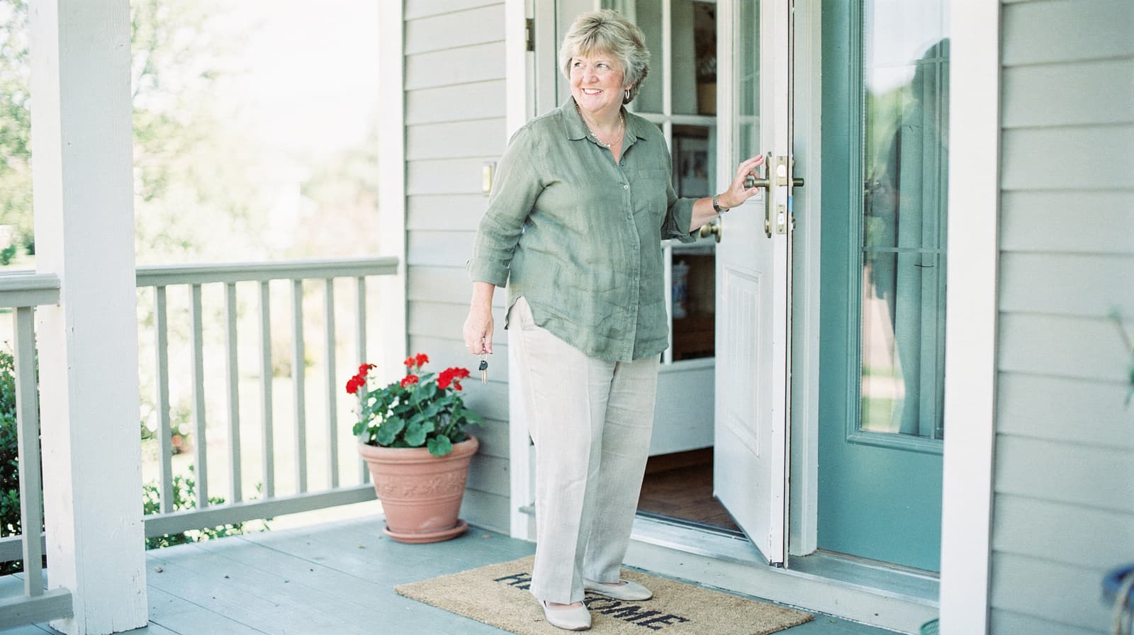 A woman in a green shirt and beige pants steps out of a house onto a porch with a potted plant.