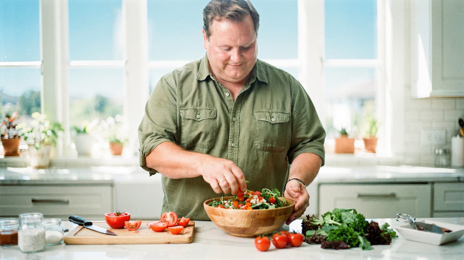 A man in a kitchen prepares a salad with fresh vegetables on a countertop.