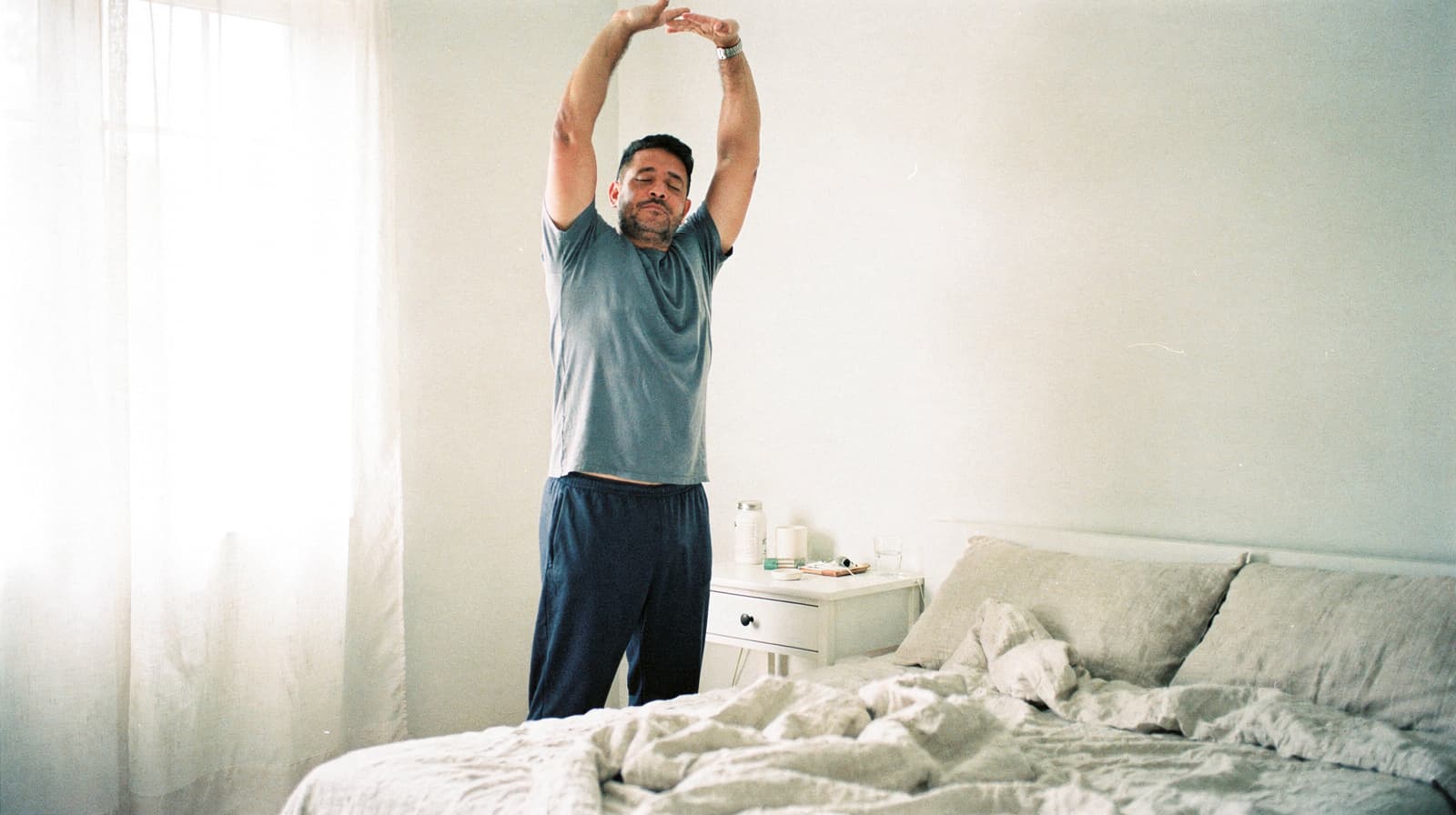 A man stretches with his arms overhead next to an unmade bed in a softly lit bedroom.
