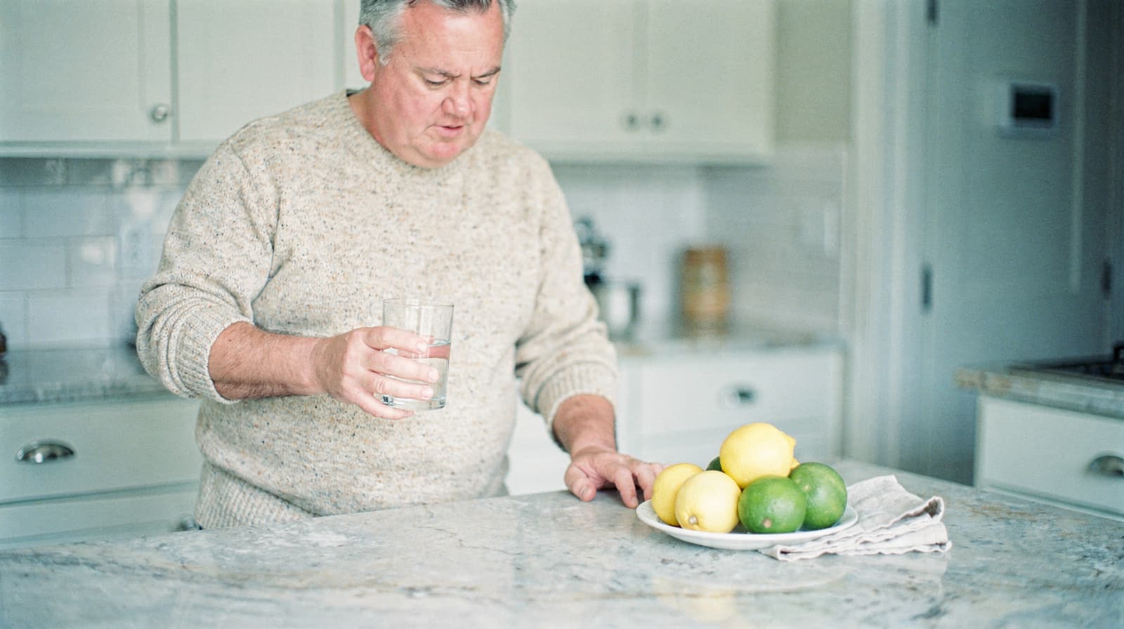 A man in a sweater holds a glass of water while standing at a kitchen counter with a plate of lemons and limes.