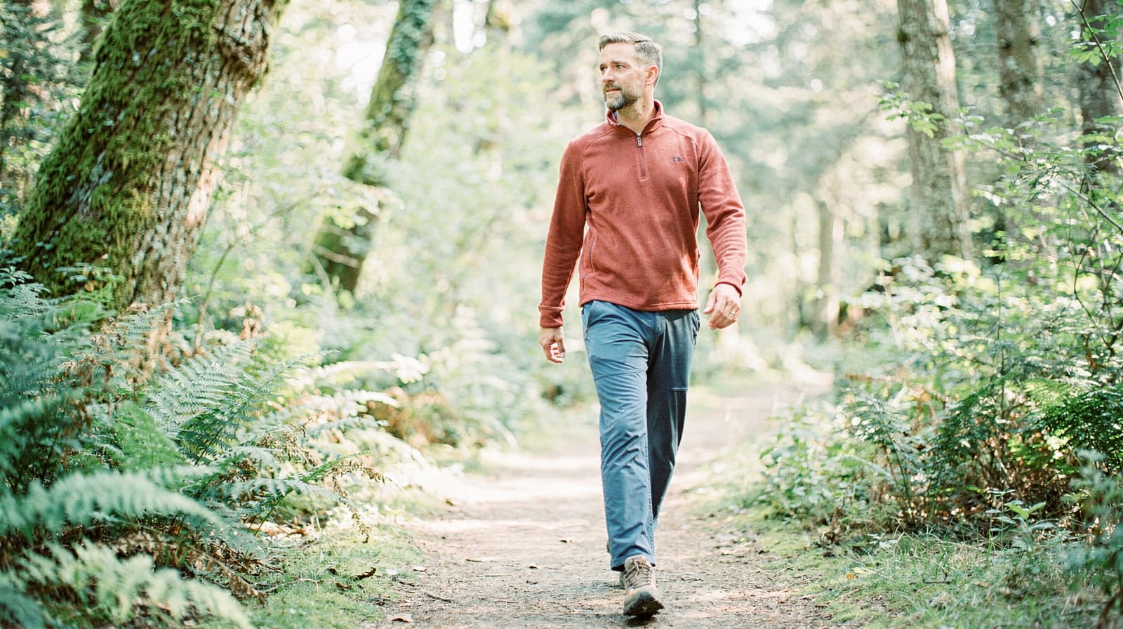 A man walks along a forest trail surrounded by trees and ferns.
