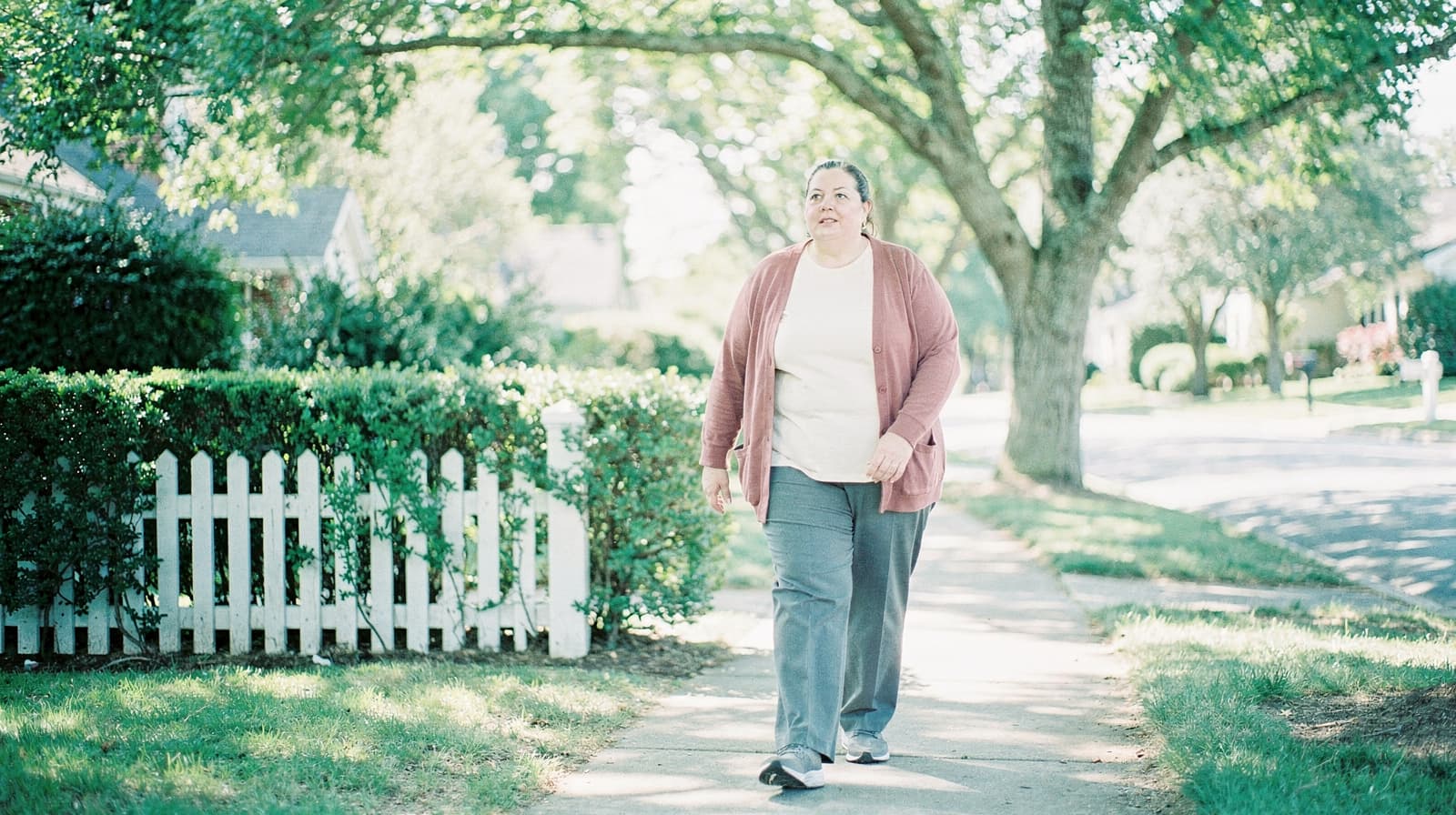 A woman walks on a sunny sidewalk in a residential neighborhood with trees and a white picket fence.
