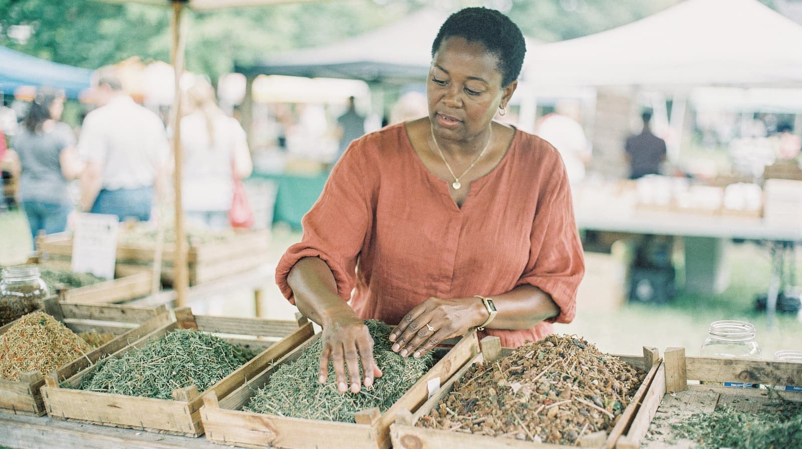 A woman in a red shirt examines dried herbs at an outdoor market stall.