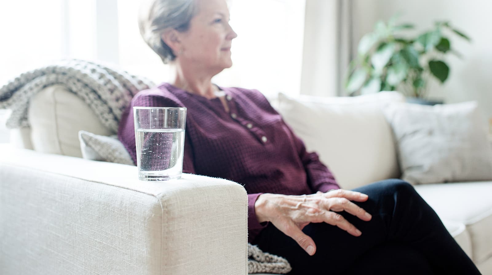 A woman sits on a sofa with a glass of water on the armrest in a bright living room.