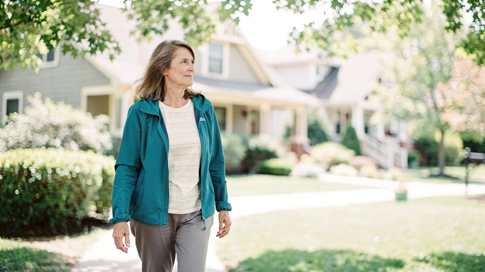 A woman in a teal jacket walks on a sunny sidewalk in a residential neighborhood with houses in the background.