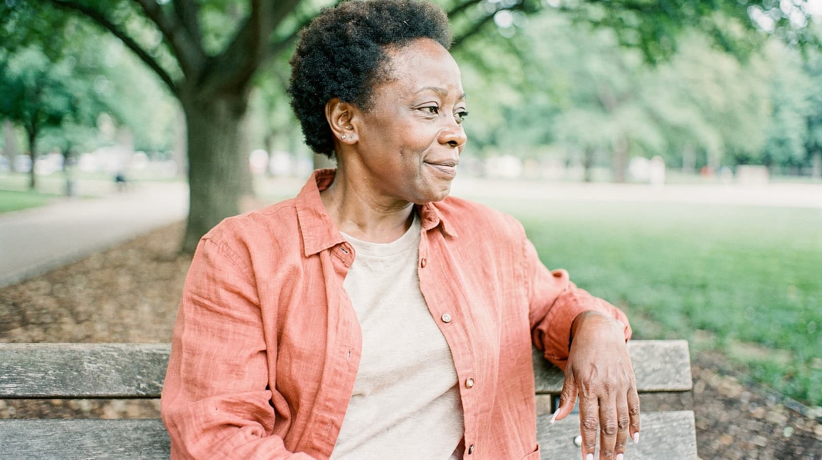 A woman sits on a park bench, looking to the side, with trees and grass in the background.