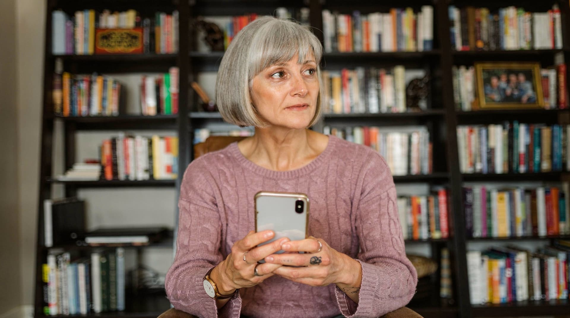 A woman with gray hair holds a smartphone while sitting in front of a bookshelf filled with books.