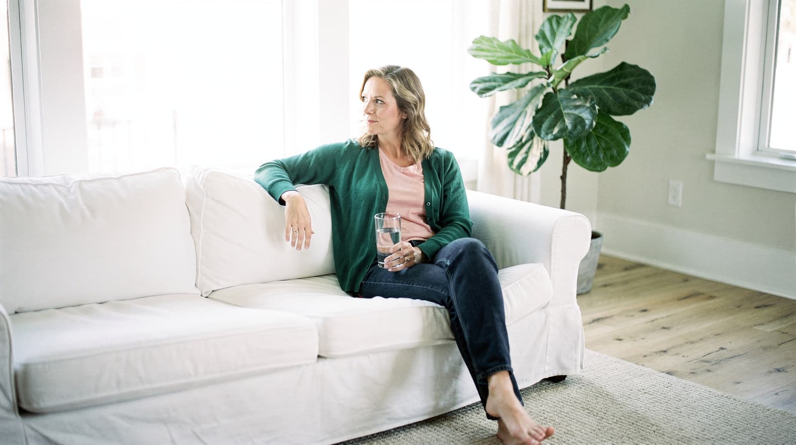 A woman sits on a white sofa holding a glass of water in a bright room with a large plant.