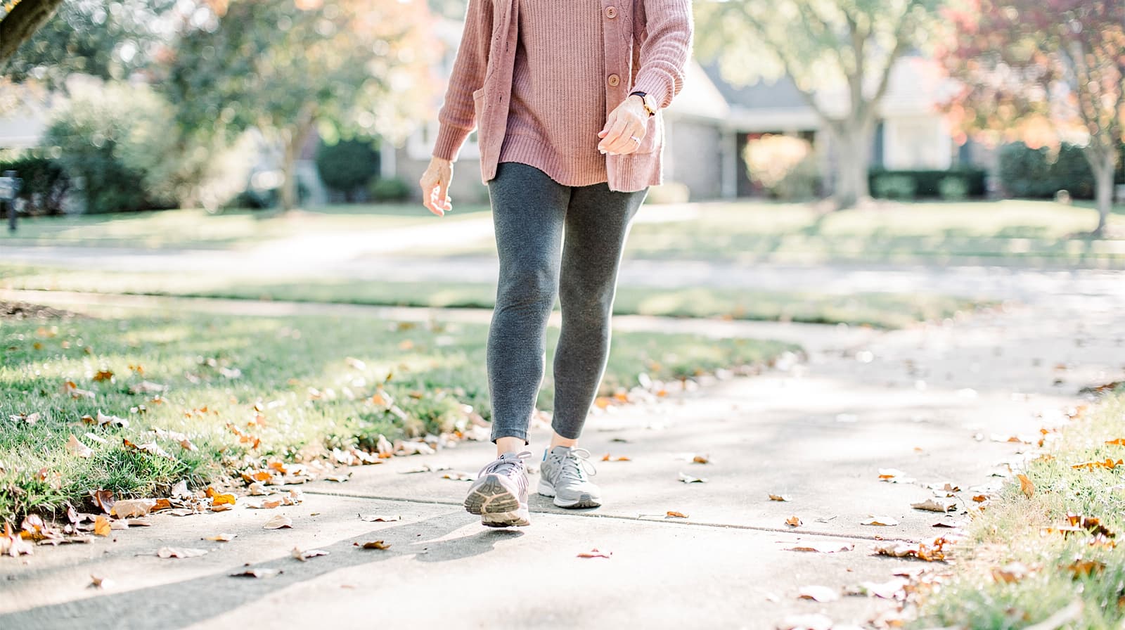 A person in a pink sweater and gray leggings walks on a leaf-strewn sidewalk in a suburban neighborhood.