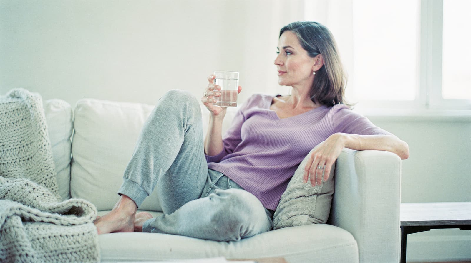 A woman in a purple sweater sits on a couch holding a glass of water, looking out a window.