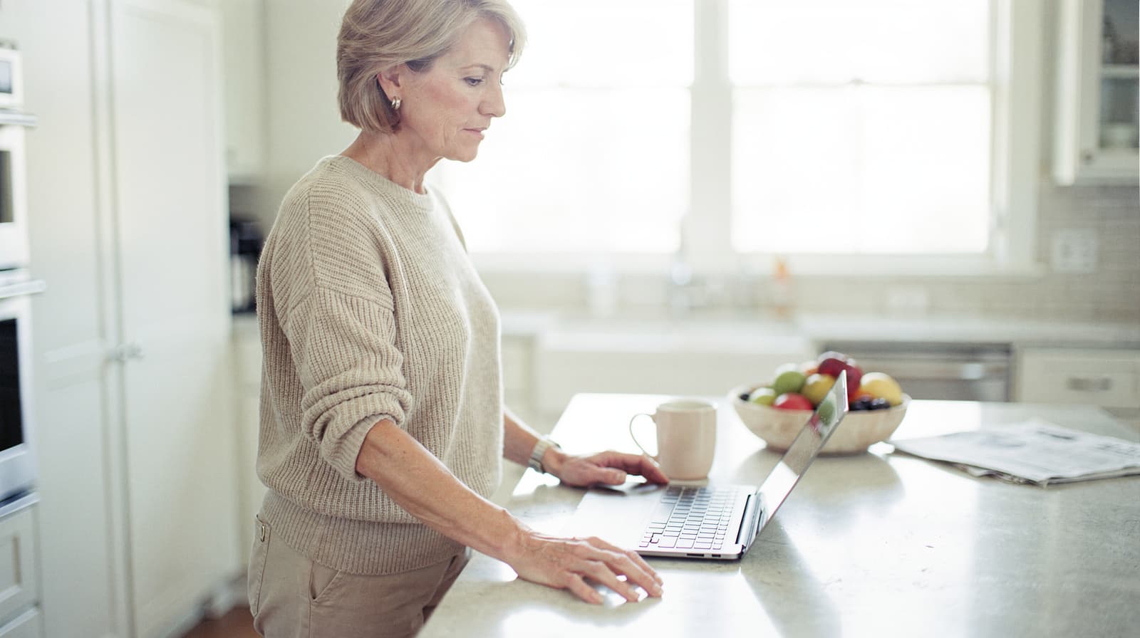 A woman stands in a kitchen using a laptop on a counter with a mug and a bowl of fruit nearby.
