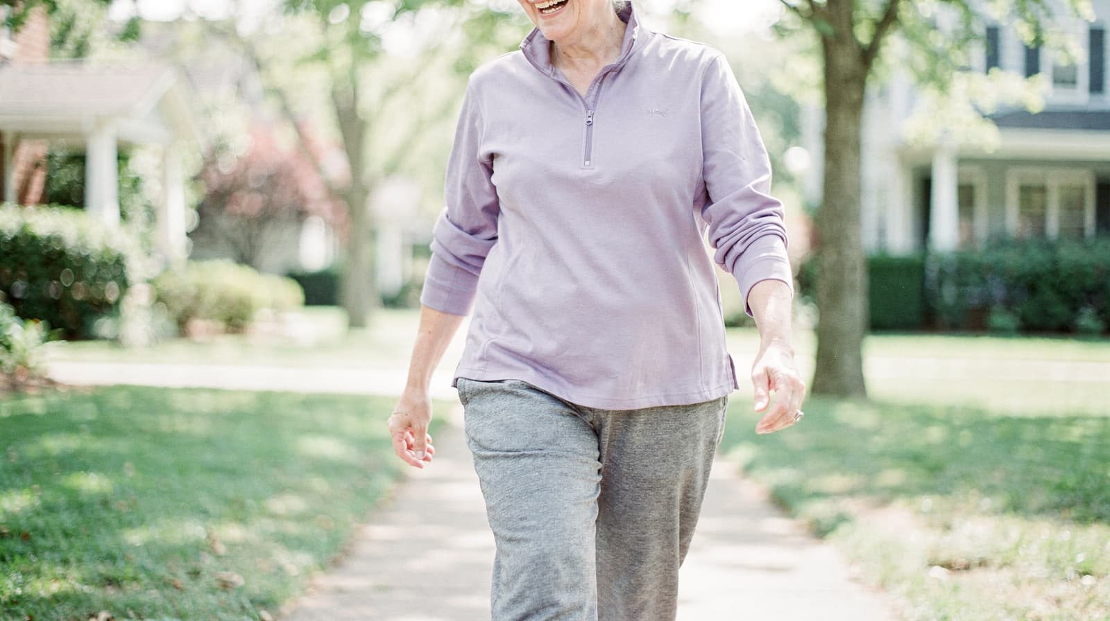 A woman in a purple top and gray pants walks on a sunny sidewalk in a residential neighborhood.